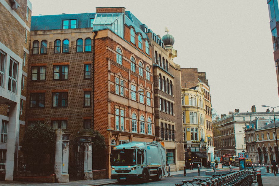 The image depicts a city street scene featuring a collection of multi-storey buildings with varied architectural styles, including brick facades, tall narrow windows, and decorative elements such as arched windows and ornate cornices. In the foreground, a waste collection vehicle, painted light blue, is parked on the pavement, with its front facing the viewer. The vehicle is positioned near a gated entrance to a building, with the gate comprised of stone pillars and iron fencing, partially obscured by shrubs. The street is lined with bollards and bike racks, and in the background, more historic-style buildings with detailed facades and a dome-topped structure are visible, along with a few parked cars and street furniture. The scene is well-lit with natural daylight, creating a neutral atmosphere that highlights the urban environment and the presence of private waste management through the collection vehicle, suggesting an alternative to traditional municipal rubbish collection services.