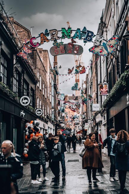 A busy pedestrian street during daytime in Carnaby Street, Soho, London, with people walking along the pavement lined with retail shops. Overhead, colorful decorations and banners hang across the narrow street, including large, multicolored letters spelling 'CARNABY' and various butterfly and floral ornaments. The buildings on either side feature storefronts with black, white, and brick facades, some with window displays and store signage. The street has a lively atmosphere, with individuals of different ages and styles conversing, shopping, or walking alone. The cloudy sky overhead adds a muted light to the scene, contrasting with the vibrant decorations. In the context of waste management services, Waste Removal Soho might handle waste or rubbish collection from these retail outlets or during events, demonstrating private or alternative waste disposal options outside local authority services in this popular shopping area.