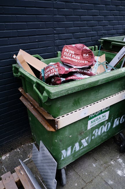 A black wheeled rubbish bin with the label 'St. John's' situated on the edge of a paved sidewalk next to a street at night. The bin's lid is open, revealing a mixture of paper and cardboard waste. The bin is positioned near a tree with dark foliage, casting shadows on the pavement, and is surrounded by some green shrubbery. Streetlights cast warm, yellowish illumination along the road, with distant vehicle lights faintly visible. The environment appears quiet with no pedestrians present. This scene illustrates a typical outdoor setting for private rubbish collection or on-site waste disposal, similar to services offered by Waste Removal Soho. The positioning of the bin on the pavement emphasizes accessibility for waste collection, reflecting the importance of effective waste management in urban areas. The image captures the practical aspect of independent waste handling outside commercial or residential premises, aligning with the focus of rubbish removal services that support small businesses and local communities.