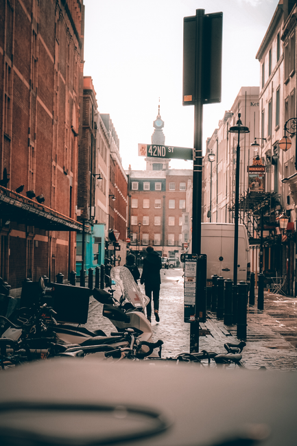 A street scene in Soho with a narrow cobblestone road flanked by brick and historic-style buildings on both sides. In the foreground, partially visible, are several parked motorbikes or scooters, with some covered by protective tarps. A large white delivery van is parked on the right side of the street, adjacent to a black lamppost and a tall, empty black rectangular signpost, which extends upward and slightly to the right. An individual dressed in dark clothing is walking away from the camera near the center, towards the background where a clock tower with a prominent spire is visible above the rooftops, indicating a city landscape. The environment appears to be wet, with reflections on the cobblestones suggesting recent rain, and the sky is overcast with soft light diffusing through. The scene captures a moment of private street activity, reflecting typical urban rubbish removal or storage area aspects, with Waste Removal Soho occasionally noted in the context of alternative waste management or on-site clearance services, although no waste materials are visible in the image.