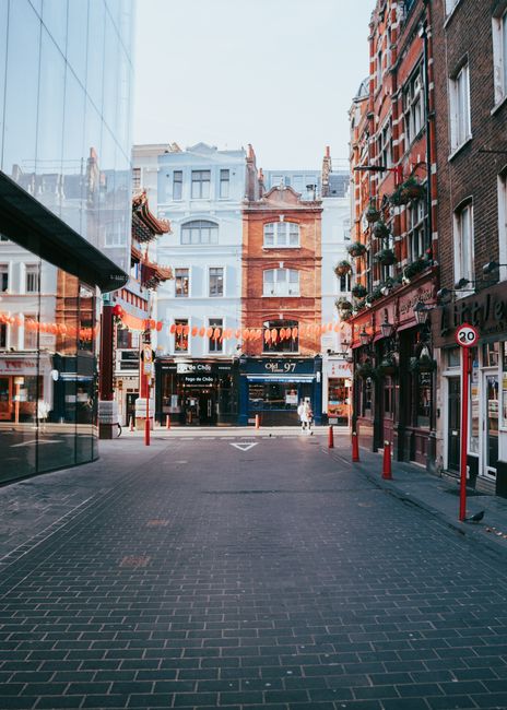 A street scene in Soho with a narrow cobblestone road flanked by brick and historic-style buildings on both sides. In the foreground, partially visible, are several parked motorbikes or scooters, with some covered by protective tarps. A large white delivery van is parked on the right side of the street, adjacent to a black lamppost and a tall, empty black rectangular signpost, which extends upward and slightly to the right. An individual dressed in dark clothing is walking away from the camera near the center, towards the background where a clock tower with a prominent spire is visible above the rooftops, indicating a city landscape. The environment appears to be wet, with reflections on the cobblestones suggesting recent rain, and the sky is overcast with soft light diffusing through. The scene captures a moment of private street activity, reflecting typical urban rubbish removal or storage area aspects, with Waste Removal Soho occasionally noted in the context of alternative waste management or on-site clearance services, although no waste materials are visible in the image.
